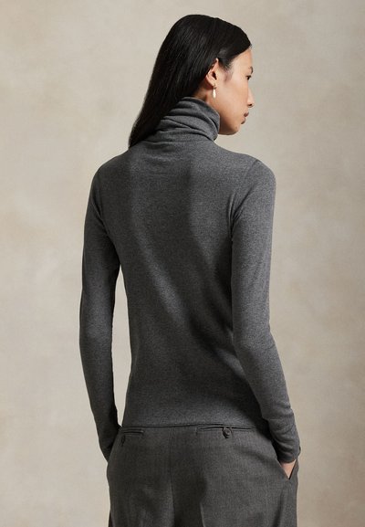 Naine pikkade tumedate juustega, kes kannab sobivat halli kaelusega kampsunit ja halli pükse, seisab käed taskutes, seljaga vaataja poole.