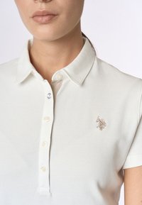 Polo shirt bianco realizzato in tessuto testurizzato. Presenta un colletto classico, quattro bottoni e un piccolo logo ricamato sul lato sinistro del petto.