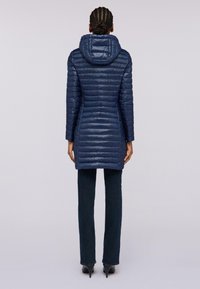Giacca puffer blu navy con cappuccio, caratterizzata da pannelli verticali trapuntati e un design di lunghezza intermedia. Indossata con jeans blu scuro e tacchi.