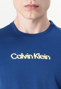Μπλε βαμβακερό t-shirt με στρογγυλή λαιμόκοψη και χαλαρή εφαρμογή, με ένα κίτρινο παραμορφωμένο λογότυπο "Calvin Klein" στο στήθος.