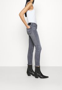 Jeans grises ajustados con una cintura media, bolsillos laterales y costuras visibles, combinados con botas negras hasta el tobillo y una camiseta sin mangas blanca.