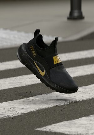 Black athletic shoe with a textured upper, gold accents, and a prominent “NIKE” logo. Features speckled sole and slip-on design.