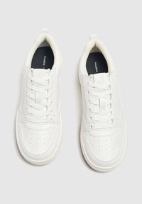Un paio di sneaker bianche basse con lacci, punte perforate e soletti neri, mostrato dall'alto su uno sfondo grigio chiaro.