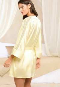 Rochie din satin galben cu accente din dantelă pe mâneci, tăietură până la genunchi și o centură legată în talie. Textură netedă, croială lejeră.