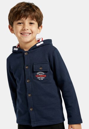 Jeune garçon souriant, portant une veste à capuche bleu marine avec des boutons et un logo sur la poche indiquant "Lee Cooper Denim Company Est. 1908".