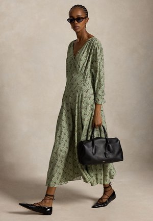 Donna con un lungo vestito verde a fantasia, occhiali da sole neri a forma di occhio di gatto, ballerine nere con lacci alla caviglia, che porta una grande borsa nera.