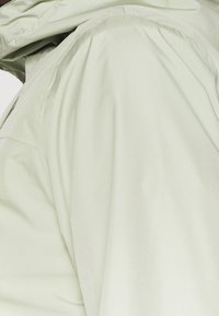 Ljusgrön jacka med en smidig, glansig yta och huva. Har subtila veck och sömmar som framhäver en modern, funktionell design.