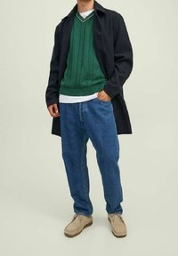 Maglione verde lavorato a maglia con motivo a treccia, indossato sopra una t-shirt bianca, sotto un cappotto scuro. I pantaloni in denim blu e le scarpe color sabbia completano il look.