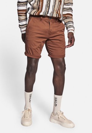 Brown cotton shorts with turned-up cuffs, paired with beige socks featuring black lettering and light-colored sneakers. Striped long-sleeve shirt.