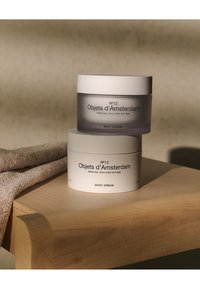 Due contenitori di prodotti per la cura del corpo impilati su una superficie di legno: un barattolo grigio opaco etichettato "SCRUB CORPO" e un barattolo bianco etichettato "CREMA CORPO."