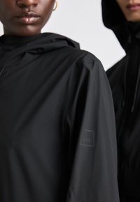 Jachetă neagră cu glugă, cu o textură netedă și o mică patch cu logo pe mânecă. Design simplu, cu un fermoar frontal și un stil minimalist.