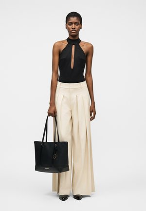 Femeie purtând un top negru fără mâneci, tip halter, pantaloni largi bej cu talie înaltă, pantofi negri cu vârf ascuțit, ținând o geantă mare neagră tip tote.