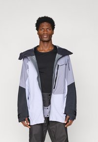 Άνδρας που φοράει ανοιχτό μωβ και μαύρο εξωτερικό μπουφάν πάνω από μαύρο μπλουζάκι και παντελόνι, στέκεται μπροστά σε λευκό φόντο.