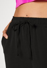 Pantaloni neri con cordoncino e tessuto testurizzato, dotati di una vita elastica e tasche laterali discrete, mostrati con un top corto rosa sopra.