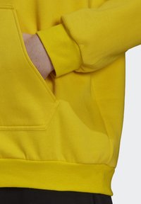 Gul sweatshirtstof med en struktureret overflade. Inkluderer en kengurulomme og ribbede manchetter. Nærbillede af lommen og ærmeområdet.