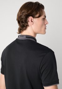 Svart polo skjorta med en grå ribbad krage, subtila sömdetaljer och en tryckt logotyp på baksidan av nacken. Placeras mot en enfärgad bakgrund.