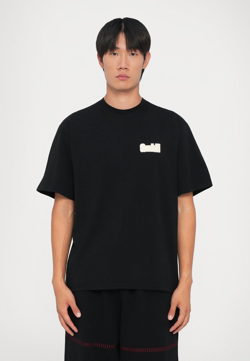 Ung mand med glat sort hår iført en løs sort T-shirt med hvidt logo på brystet og sorte shorts med røde detaljer.