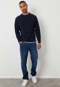 Felpa blu navy con collo rotondo, maniche lunghe raglan, abbinata a jeans in denim blu. Il modello indossa sneaker bianche e una maglietta bianca sotto.