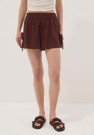 Donna che indossa pantaloncini marroni con vita elasticizzata e laccetti laterali, top corto bianco e sandali marroni con fibbie e borchie metalliche.