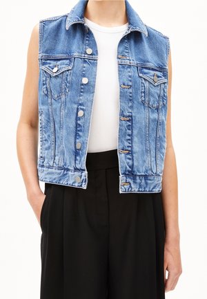 Persoon draagt een mouwloos blauwe spijkervest met zilveren knopen over een wit topje en losse zwarte broek, handen in de zakken.