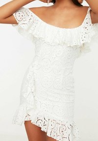 Valkoinen olkapäiltä avattu mekko, jossa on rypytysmyssy, pitsi- ja ympyräkuvioita, sekä kerrokselliset rypytykset helman kohdalla.