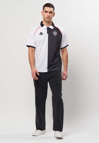 Homme debout, portant un polo moitié noir, moitié blanc avec des logos, un pantalon noir et des baskets blanches sur un fond uni clair.
