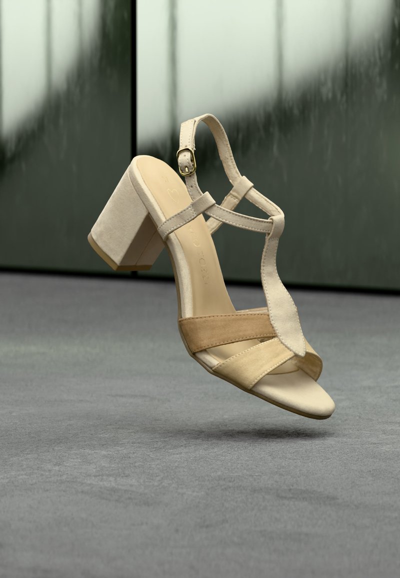 Μπεζ σουέντ και δερμάτινο ψηλοτάκουνο σανδάλι με λουράκι αστραγάλου και πόρπη, απεικονίζεται στον αέρα πάνω από γκρι πάτωμα και πράσινο υπόβαθρο.