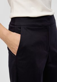 Zwarte op maat gemaakte broek met zijzakken, voorzien van een gladde textuur en een aangesloten ontwerp. Een witte top is gedeeltelijk zichtbaar boven de tailleband.