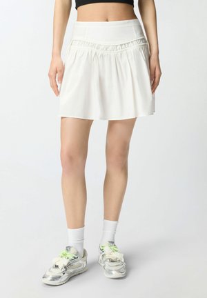 Donna che indossa una minigonna plissettata bianca, calzini bianchi e scarpe da ginnastica argento con lacci verde neon, in piedi contro uno sfondo uniforme.