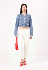 Blauwe cropped jas van gestructureerde stof met voorzakken, gecombineerd met witte jeans. Houdt een rode tas vast en draagt rode schoenen met gespen.