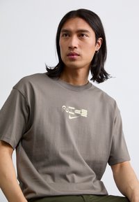 En ung mand med langt hår har en grå t-shirt med et stiliseret grafisk logo på brystet, siddende mod en ensfarvet baggrund.