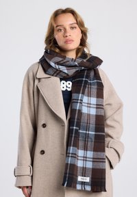 Karierter Schal in Braun, Blau und Weiß, aus weichem Stoff, um den Hals gewickelt, kombiniert mit einem beigen Wollmantel.