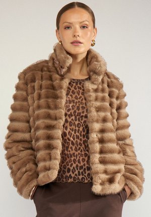 Woman wearing a brown striped faux fur jacket over a leopard print top with hands in dark brown pants pockets against a plain background.