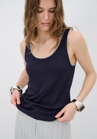 Tmavě modrý žebrovaný tílko s kontrastním světlým prošíváním podél výstřihu. Model má na sobě stříbrné náramky a šedé proužkované kalhoty.