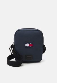 Granatowa torebka typu crossbody z zapięciem na zamek, z naszywką z logo w kolorach czerwonym, białym i niebieskim. Regulowany czarny pasek i gładka faktura.