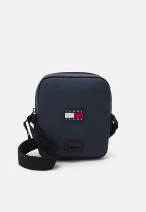 Borsa a tracolla blu navy con chiusura a zip, dotata di una toppa con logo in rosso, bianco e blu. Spallaccio nero regolabile e superficie liscia.