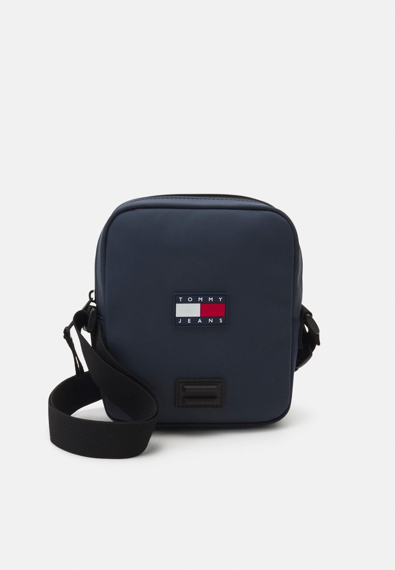 Borsa a tracolla blu navy con chiusura a zip, dotata di una toppa con logo in rosso, bianco e blu. Spallaccio nero regolabile e superficie liscia.