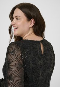 Robe noire avec des motifs de sequins intricats, des manches longues transparentes et un dos en clé, avec un tissu texturé et un design élégant.