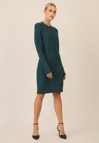 Teal kabelstrik kjole med lange ærmer og ribbede manchetter, med en tætsiddende silhuet og knælang kant. Sorte højhælede sko.