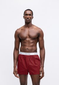 Raudoni dryžuoti apatiniai šortai su balta Calvin Klein juosta. Tekstūra lygi, su geometriniu raštu kontrastinga spalva.