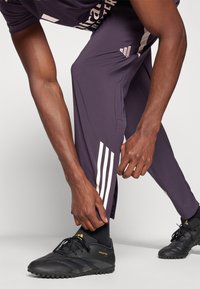 Schwarze Sportschuhe mit strukturierten Sohlen; enge dunkelviolette Hosen mit weißen Streifen an den Seiten; Hände, die den Hosenumschlag anpassen.