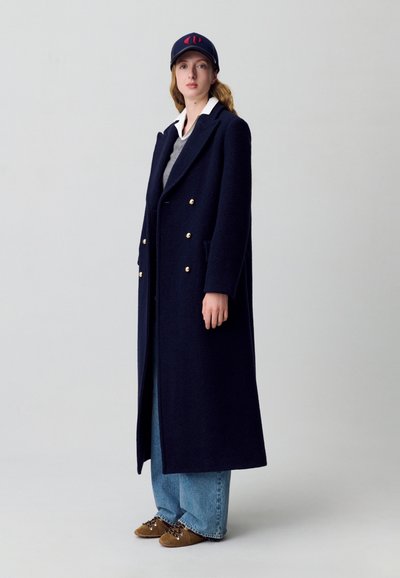 Cappotto doppiopetto in misto lana blu navy con bottoni dorati, dalla vestibilità oversize, abbinato a un maglione grigio, jeans blu e scarpe stringate marroni.