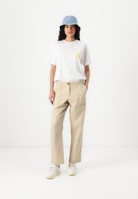T-shirt in cotone bianco con testo grafico giallo, pantaloni beige a gamba larga con tasche frontali e cappello bucket blu. Sneakers casual completano il look.