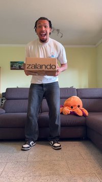 Brun pappeske med "zalando" trykt i svart, holdt av en person kledd i hvit t-skjorte og grå jeans. Orange plysjblekksprut på sofaen.