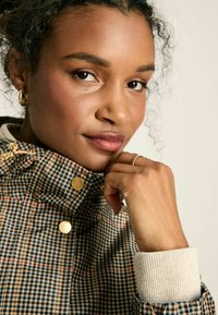 Pārbaudītas auduma jaka ar zeltainām pogām, ar augstu apkakli un ribotu aproci. Krāsas ir bēša, jūras zila un bordo.