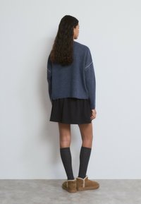 Mujer con cabello largo y rizado, vistiendo un suéter azul, falda negra, calcetas oscuras hasta la rodilla y botas cortas color beige forradas de piel, parada contra una pared blanca.