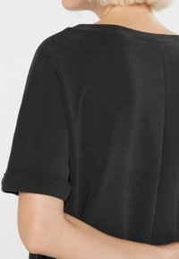 Woman with short blonde hair wearing a black short-sleeve top with rolled cuffs, shown from the back and side against a white background.