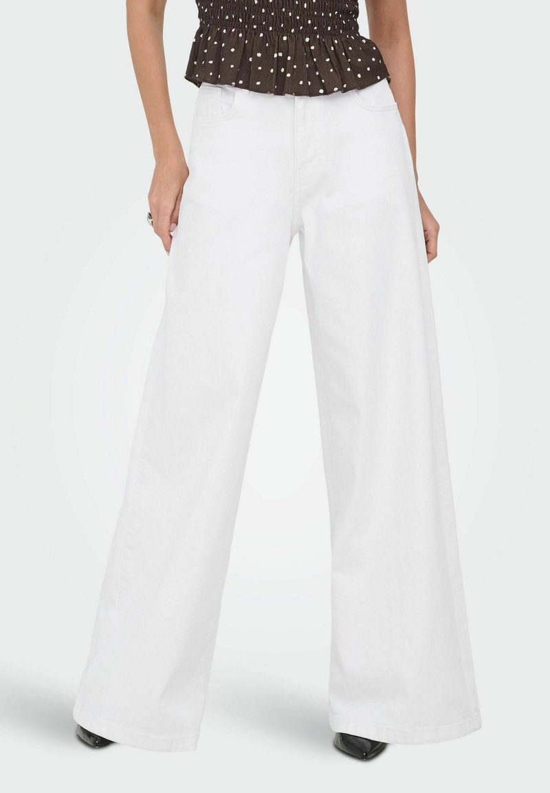 Femme portant un pantalon blanc à jambes larges assorti d'un haut noir à pois blancs, se tenant debout avec les mains détendues le long du corps.