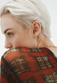 Pendiente en forma de U de color dorado en la oreja; blusa de patrón multicolor en tonos de rojo, naranja y amarillo. Cabello corto y rubio claro.