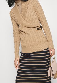 Sweter w warkocze w beżowym kolorze z szerokim kołnierzem i paskiem z boku; skomponowany z ciemną spódnicą w paski z beżowymi paskami.